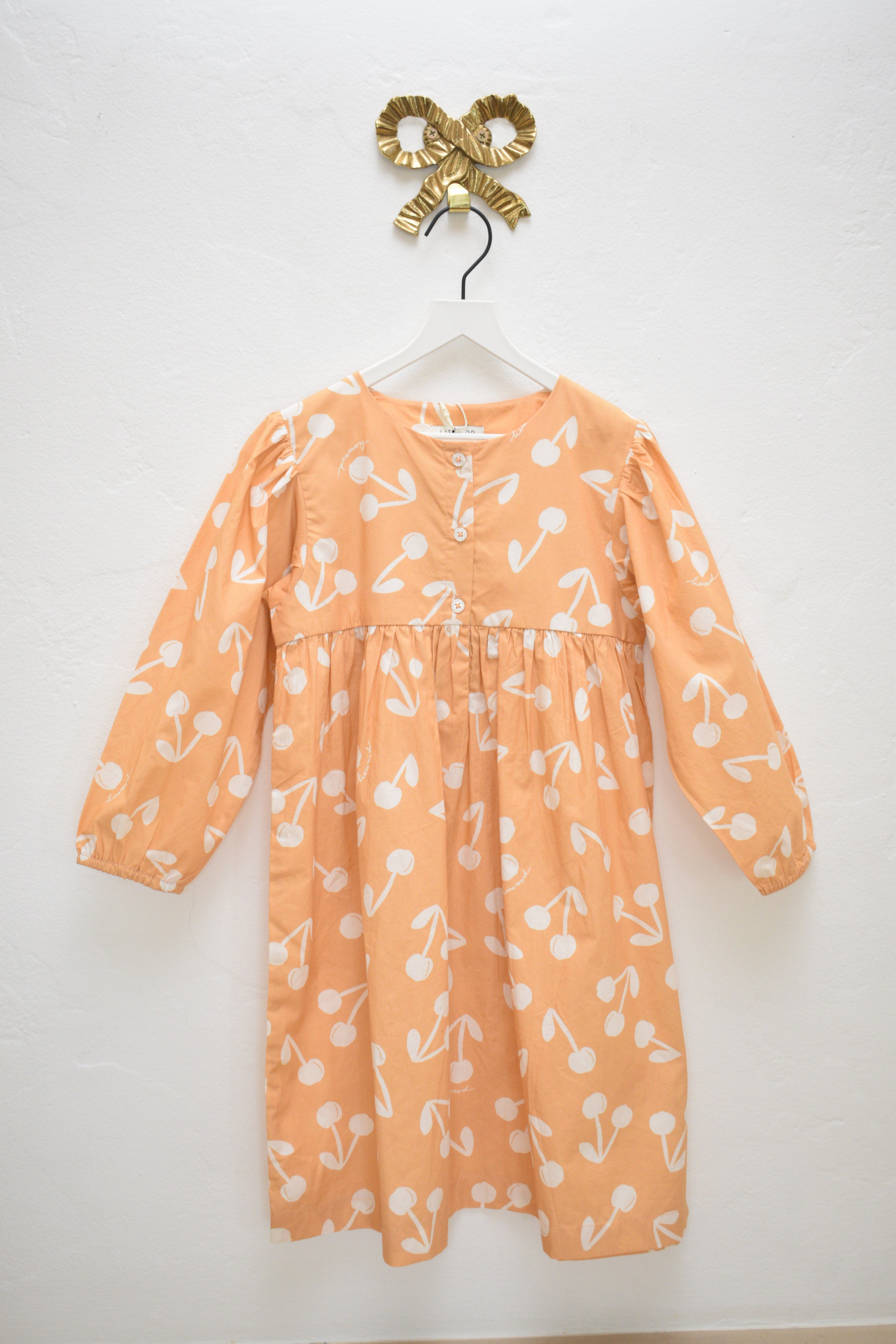 Liewood Ariana peach dress with ivory cherry print, size 4 years, long sleeves and button placket, hanging on a white hanger against a neutral wall.
