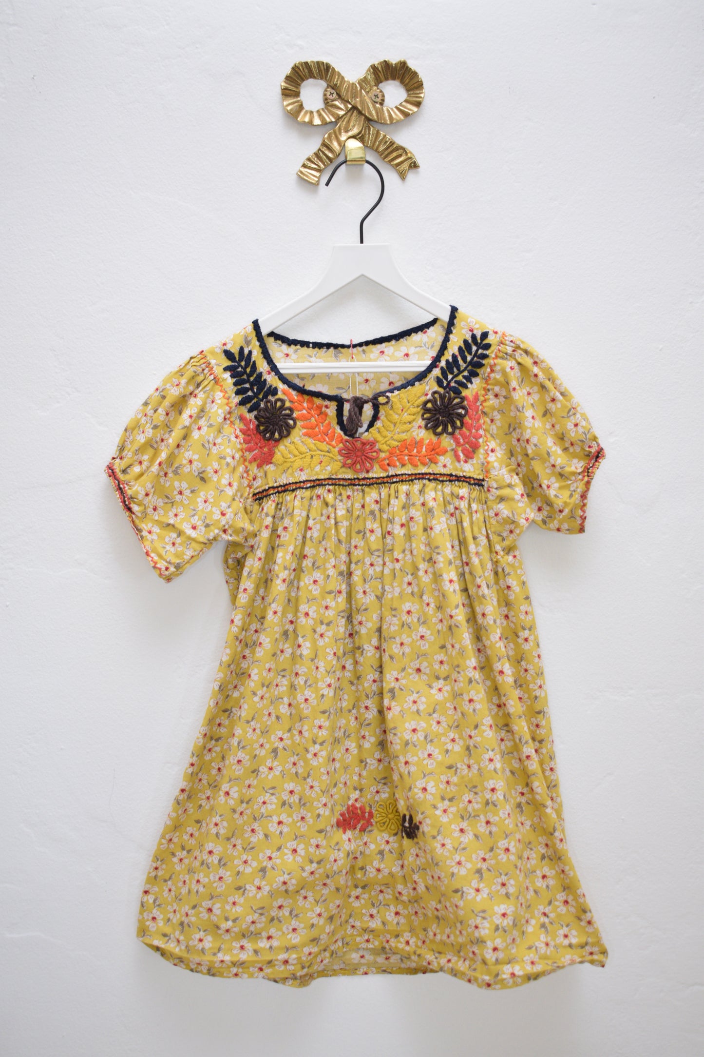 ASA ‘Garden in Fall’ marigold dress with multicolor floral print and embroidered yoke, tie neckline, size 2–3 years, hanging on a white hanger against a neutral wall.