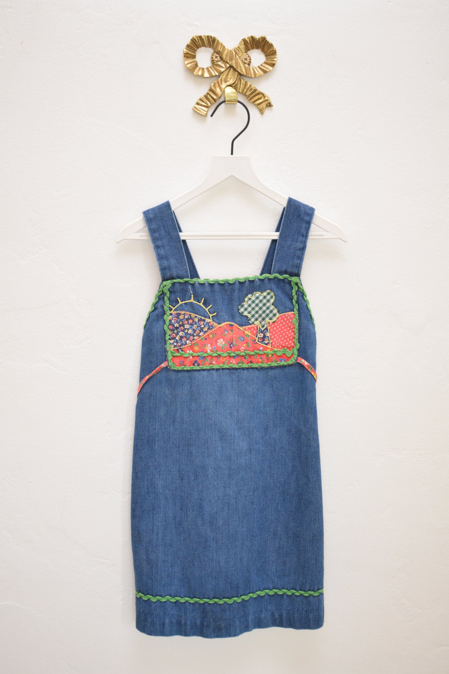 Vintage girls’ Rosebud Duds denim dress, size 5, with red calico yoke, green ric-rac trim, smocked back and tie, hanging on a white hanger.
