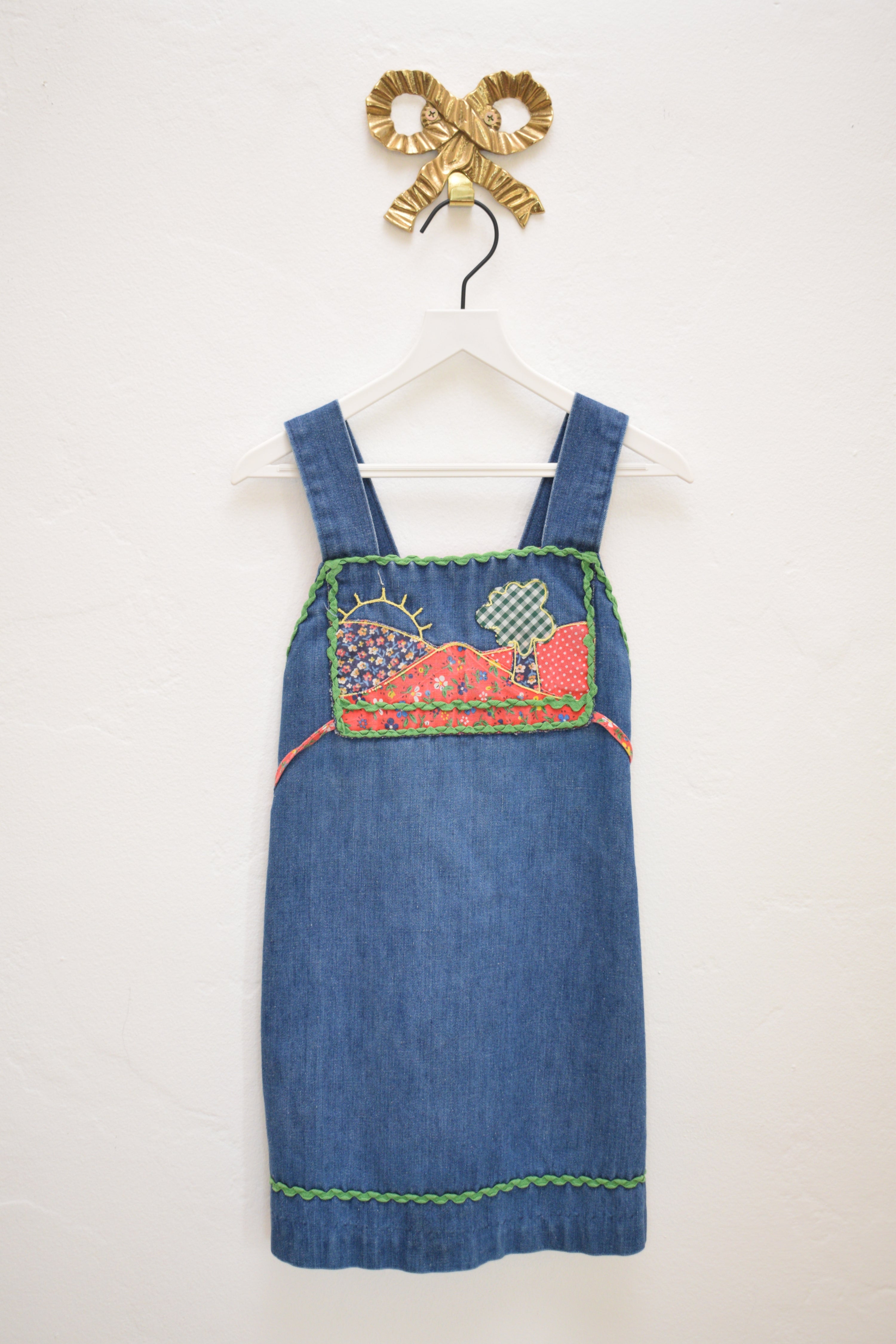 Vintage girls’ Rosebud Duds denim dress, size 5, with red calico yoke, green ric-rac trim, smocked back and tie, hanging on a white hanger.
