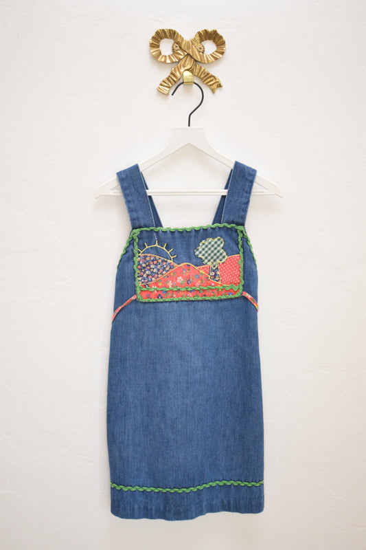 Vintage girls’ Rosebud Duds denim dress, size 5, with red calico yoke, green ric-rac trim, smocked back and tie, hanging on a white hanger.
