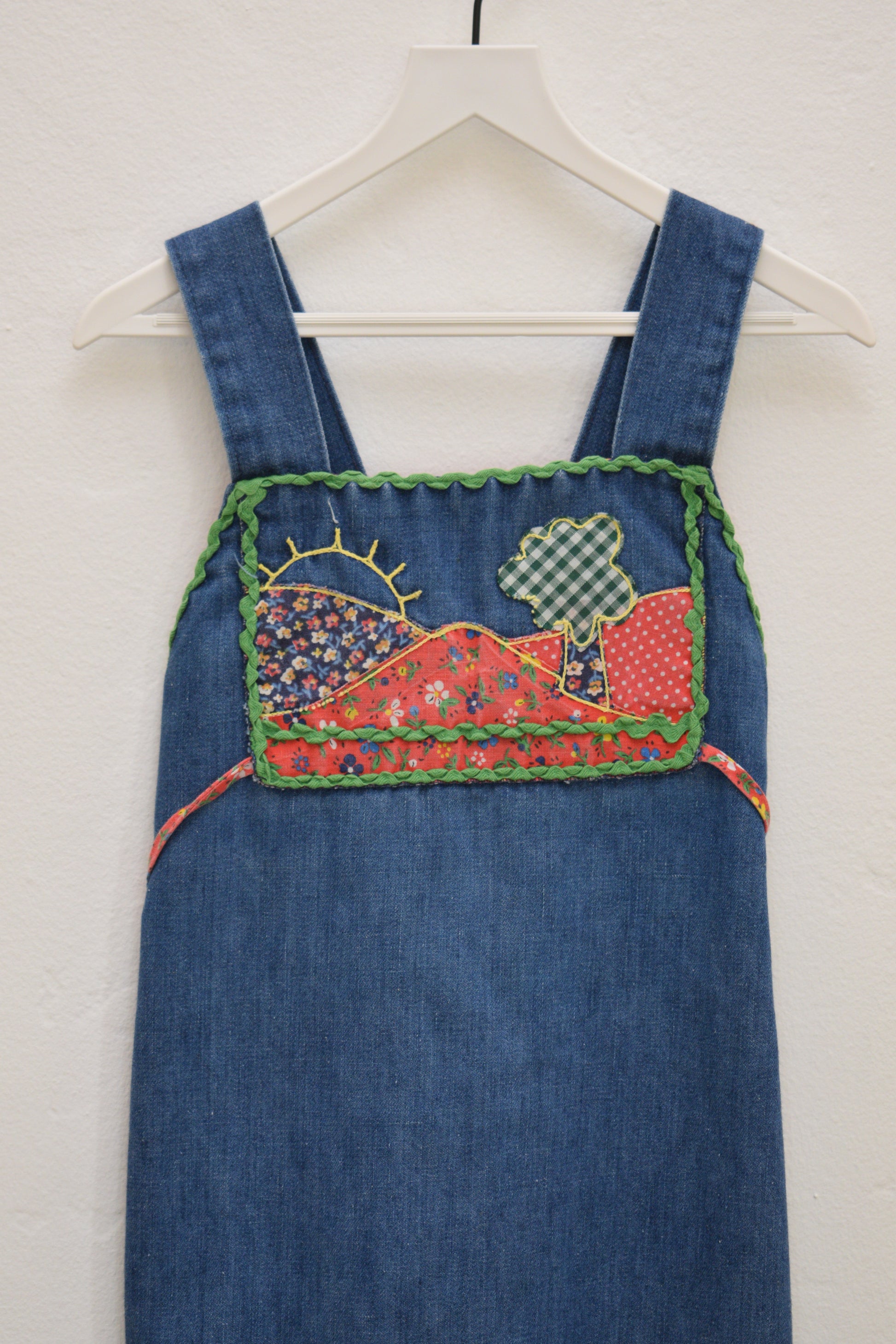 Vintage girls’ Rosebud Duds denim dress, size 5, with red calico yoke, green ric-rac trim, smocked back and tie, hanging on a white hanger.