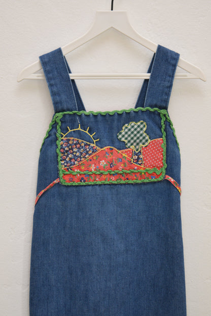 Vintage girls’ Rosebud Duds denim dress, size 5, with red calico yoke, green ric-rac trim, smocked back and tie, hanging on a white hanger.