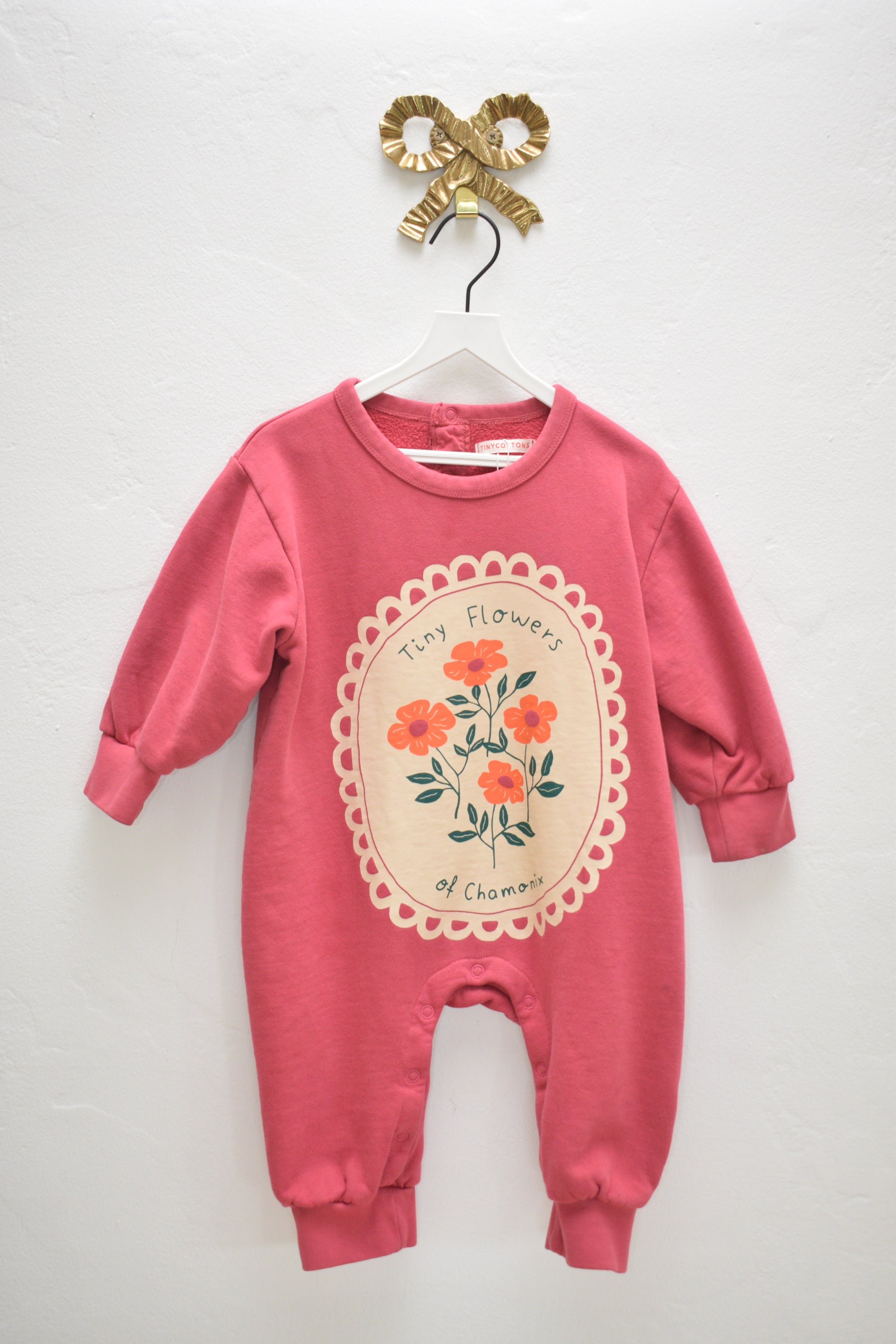 Tinycottons rose pink fleece sweat jumpsuit with ‘Tiny Flowers of Chamonix’ floral cameo print, size 24 months, on a white hanger against a neutral wall.